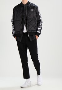 Chaqueta bomber acolchada negra con el logo blanco de adidas y tres rayas blancas en las mangas, combinada con pantalones negros y zapatillas blancas.