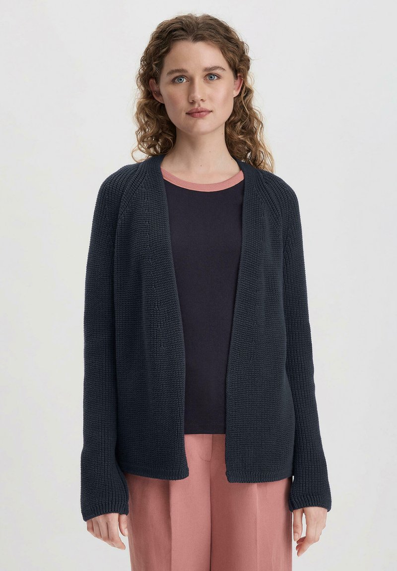 Sininen neulevillatakki, jossa on teksturoitu kuvio, avoin etuosa ja pitkät hihat. Varnattu tumman yläosan päällä, jossa on vaaleanpunainen kaula-aukko.