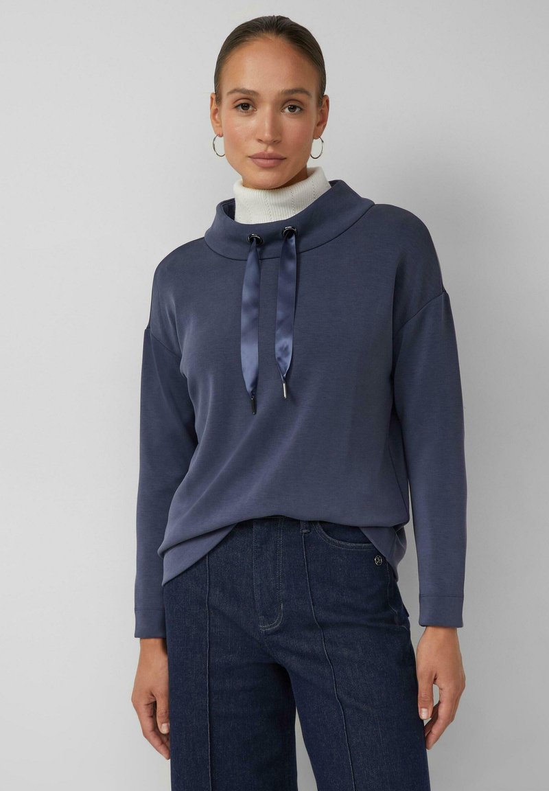 Marineblauer Pullover mit hohem Rollkragen, ausgestattet mit Satin-Kordeln und einem lockeren Schnitt. Getragen über hoch taillierter dunkler Jeans.