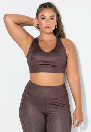 Mujer con pelo largo, con un top corto deportivo de estampado de leopardo marrón y leggings, de pie con un brazo levantado detrás de la cabeza.