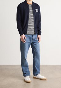 Marinblå bomberjacka med vit "HUGO" logotyp, randig crew-neck t-shirt, ljusblå jeans och vita sneakers med gummisåler.