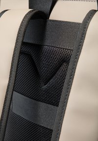 Rygsækdetalje med polstret mesh-panel, sorte nylonremme og ydre i beige. Indeholder et trekantet accentdesign med struktureret finish.