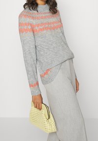 Jasnoszary dziergany sweter z okrągłym dekoltem, ozdobiony koralowymi i złotymi geometrycznymi wzorami na karczku. Trzymana jest teksturowana żółta torebka.