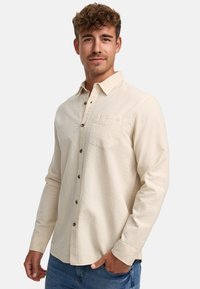 Chemise beige à manches longues en tissu texturé, avec un devant à boutons, un col, une poche poitrine unique et des détails de couture subtils.