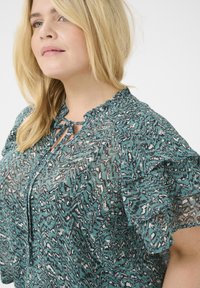 Blusa fluida en color teal con un escote y mangas con volantes. Presenta un patrón ondulado en blanco y negro. Tejido ligero y semitranslúcido.