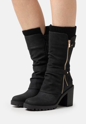 Black mid-calf heeled boots with gold side zippers, strap detailing, and ribbed knit cuffs worn on bare legs against a plain background.