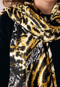 Person trägt ein schwarzes Oberteil und einen gelb-schwarz-weißen Schal mit Tiermuster um den Hals gewickelt.