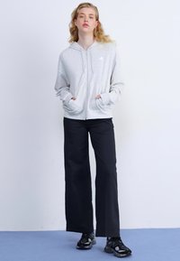 Un sweat à capuche zippé gris clair avec une capuche, des poches avant et des poignets côtelés, associé à un pantalon noir à jambe large et des baskets noires.