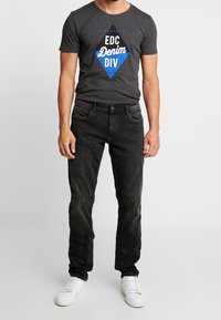 Camiseta gris con diseño gráfico en azul y negro, combinada con jeans oscuros y zapatillas blancas. El ajuste es casual con una silueta clásica.
