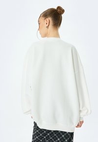 Sudadera blanca de gran tamaño hecha de tela suave, con hombros caídos, puños acanalados y cuello redondo. Llevada sobre una falda de cuadros.