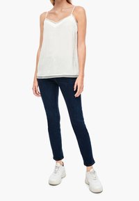 Witte mouwloze top met kantafwerking, textuurstof; gecombineerd met donkerblauwe skinny jeans en witte sneakers, getoond op een staande model.