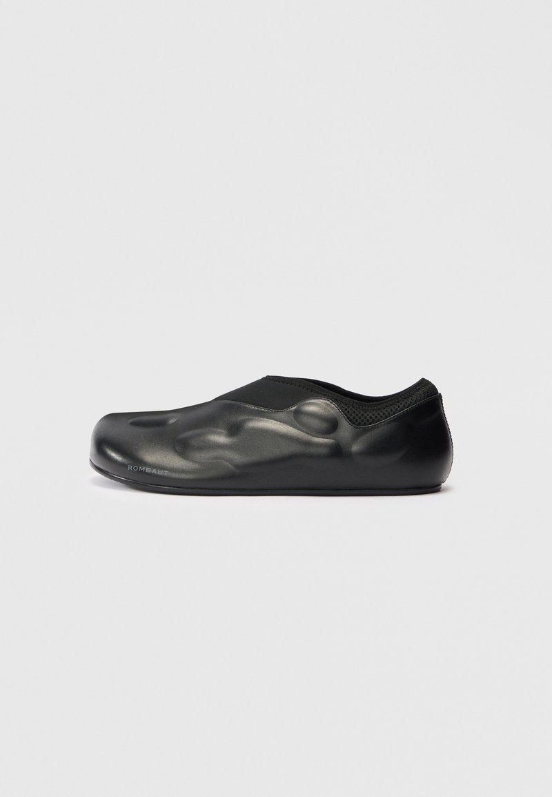 Scarpa nera slip-on con superficie liscia e texturizzata, caratterizzata da una finitura opaca e un design a profilo basso. Accenti in rete lungo il colletto.