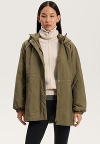 Vihreä oliivinvärinen tikattu takki, jossa on huppu, napit ja joustavat hihansuut. Päällä vaaleanbeige poolopaita.