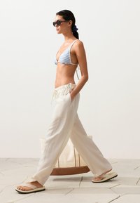 Femme marchant en pantalon blanc à cordon, haut de bikini rayé bleu et blanc, sandales blanches et lunettes de soleil foncées, portant un grand sac fourre-tout beige.