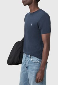 Námorně modré tričko s krátkým rukávem vyrobené z bavlny, s malým vyšitým logem na hrudi. N osené s světle modrými džíny a černou taškou.