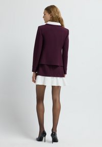 Blazer bordeaux avec col blanc, associé à une jupe bordeaux et un ourlet à volants blancs. Des escarpins noirs et des collants transparents complètent le look.