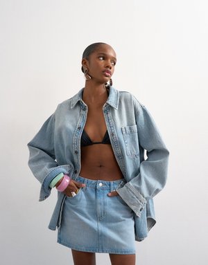 Mujer con chaqueta y falda de mezclilla azul claro de gran tamaño, con top de bikini negro, complementada con pendientes de aro y pulseras coloridas.