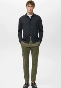 Młody mężczyzna z kręconymi włosami, ubrany w ciemnoszary rozpinany sweter, biały podkoszulek, oliwkowozielone spodnie i czarne mokasyny, stoi z rękami w kieszeniach.