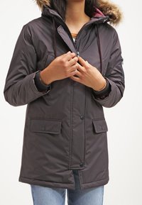 Dark grey insulated parka with a faux fur hood, front zip, and snap pockets. Features a drawstring waist and contrasting inner lining.