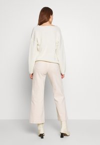 Femme portant un pull tricoté blanc, un pantalon large beige clair et des bottines à talons crème, debout face à un mur gris clair uni.