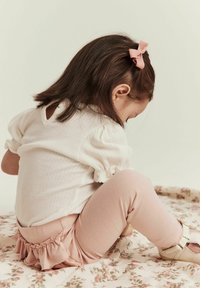 Chemise blanche à côtes avec des manches bouffantes, leggings roses avec volant à la taille, et chaussures beiges, assise sur un tissu à motif floral.