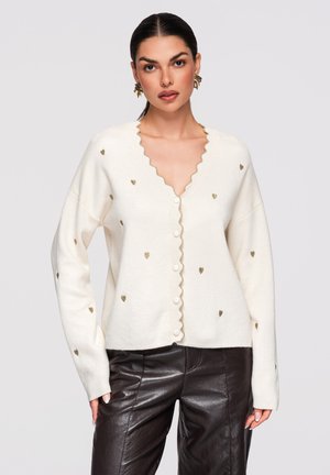 Kvinde iført en hvid knaplukket cardigan med små gyldne hjerter og bølgekant, kombineret med sorte læderbukser og gyldne øreringe.