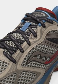 Κοντινή λήψη παπουτσιού τρεξίματος Saucony Guide 7 που δείχνει μπεζ πλέγμα στο πάνω μέρος, μαύρα κορδόνια και κόκκινες και μπλε λεπτομέρειες.