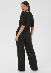 Donna che indossa un top corto a maniche corte nero con paillettes e pantaloni larghi abbinati, in piedi con la schiena rivolta, che guarda oltre la spalla.