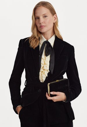 Schwarzer Samtblazer mit gebundenem Taillengürtel, darunter eine weiße Rüschbluse und eine kleine schwarze Clutch mit goldenen Akzenten. Glatte Textur und taillierte Passform.