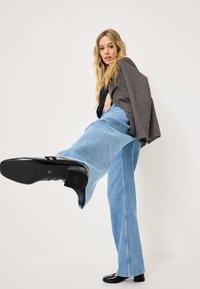 Jeans en denim bleu clair à jambes larges associés à un blazer gris foncé. Le mannequin porte des chaussures noires à finition brillante.