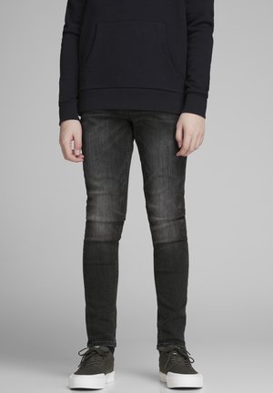 Personne portant un jean skinny noir délavé, un sweat à capuche noir et des baskets vert foncé avec des semelles blanches, debout sur un fond gris.