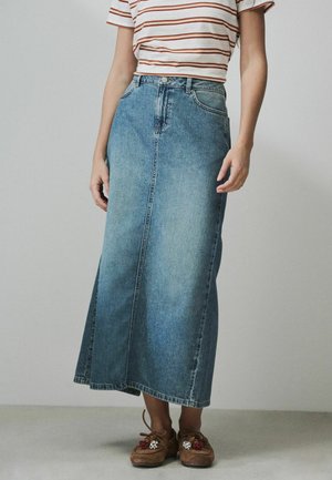 Frau trägt einen hoch taillierten, ausgewaschenen blauen Denim-Midirock, ein gestreiftes Kurzarmshirt und braune Mokassin-Schuhe mit Blumenverzierung.