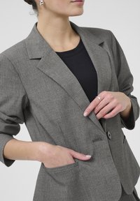 Šedý pruhovaný blazer s klopovým límcem, s jedním knoflíkem a předními kapsami, nošený přes černý top. Hladká textura látky.