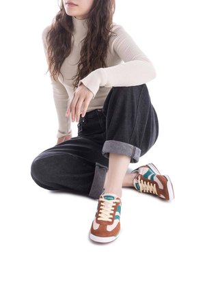Zapatillas de ante marrón y verde azulado con suela blanca, cordones beige y logo verde. El modelo lleva jeans oscuros con los dobladillos remangados.