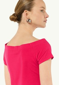 Różowa bluzka z odkrytymi ramionami i krótkimi rękawami, charakteryzująca się gładką fakturą i starannym przeszyciem. Modelka nosi duże, srebrne okrągłe kolczyki.