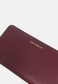 Portafoglio in pelle bordeaux con superficie testurizzata. Presenta un logo dorato, un design elegante e bordi cuciti. Forma rettangolare compatta.