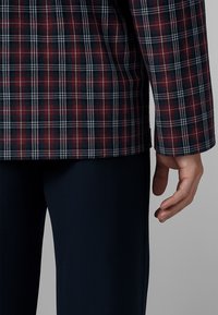 Persona che indossa una giacca a quadri rosso scuro, nera e bianca con pantaloni blu navy, mano rilassata lungo il fianco contro uno sfondo neutro.