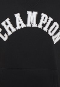 Černá látka s velkými bílými vyšívanými písmeny, které tvoří nápis "CHAMPION" v obloukovém tvaru přes oblast hrudníku.