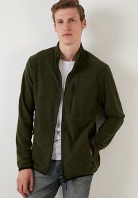 Giacca in pile verde scuro con zip frontale, colletto alto e tasca sul petto. Presenta dettagli in zip neri e tasche laterali.