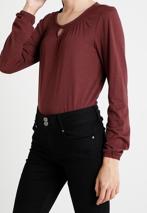 Femme portant un haut bordeaux à manches longues avec un décolleté en trou de serrure et un pantalon noir taille haute, montrant le haut du corps et le bras droit.