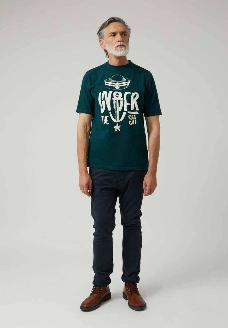 Camiseta de algodón en verde azulado oscuro con un diseño gráfico blanco de un ancla y texto. Combinada con jeans oscuros y botas marrones con cordones.