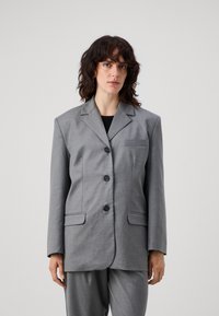Grå, overdimensjonert blazer med slått krage, fire svarte knapper og to frontlommer. Glatt stoff med minimal tekstur, kombinert med grå bukser.