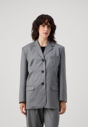 Grauer, übergroßer Blazer mit Reverskragen, vier schwarzen Knöpfen und zwei Taschen auf der Vorderseite. Glattes Material mit minimaler Textur, kombiniert mit grauen Hosen.