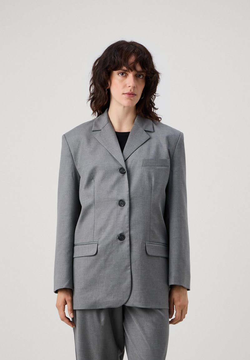 Grå, overdimensjonert blazer med slått krage, fire svarte knapper og to frontlommer. Glatt stoff med minimal tekstur, kombinert med grå bukser.