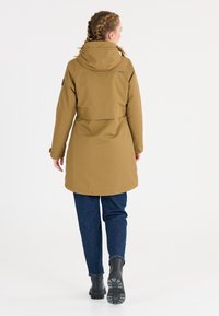 Lång brun vattentät jacka med huva, försedd med en bakre ventil, justerbara ärmar och en mjuk yta. Kombinerad med blå denimbyxor och svarta stövlar.