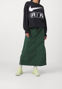 Svart sweatshirt med vitt Nike-logotyp, kombinerad med en grön maxikjol med dragsko i midjan och sidfickor. Ljusgula sneakers.