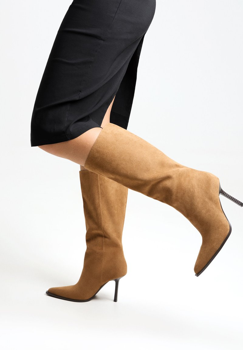 Bottes en suède beige jusqu'aux genoux avec un bout pointu et un talon aiguille. La jambe est partiellement visible, portant une jupe noire fendue.
