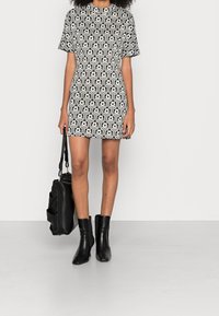 Schwarz-weiß gemustertes kurzärmeliges Kleid mit hohem Ausschnitt, A-Linie-Form, kombiniert mit schwarzen Knöchel-Stiefeln und einer schwarzen Handtasche.
