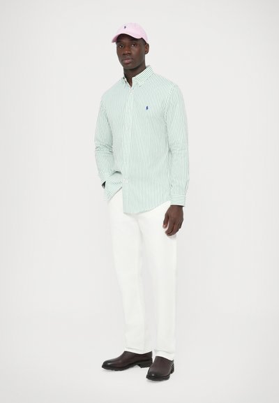 Zeleno srajco z gumbi s črtami, bele hlače in roza kapa. Na srajci je majhen moder logotip, čevlji pa so temni z teksturirano površino.