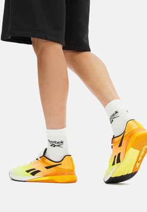 Buty sportowe Reebok z jasnosżółtą i pomarańczową cholewką, czarnymi akcentami, białymi sznurowadłami oraz teksturowaną gumową podeszwą. Krótkie czarne spodenki i białe skarpetki.